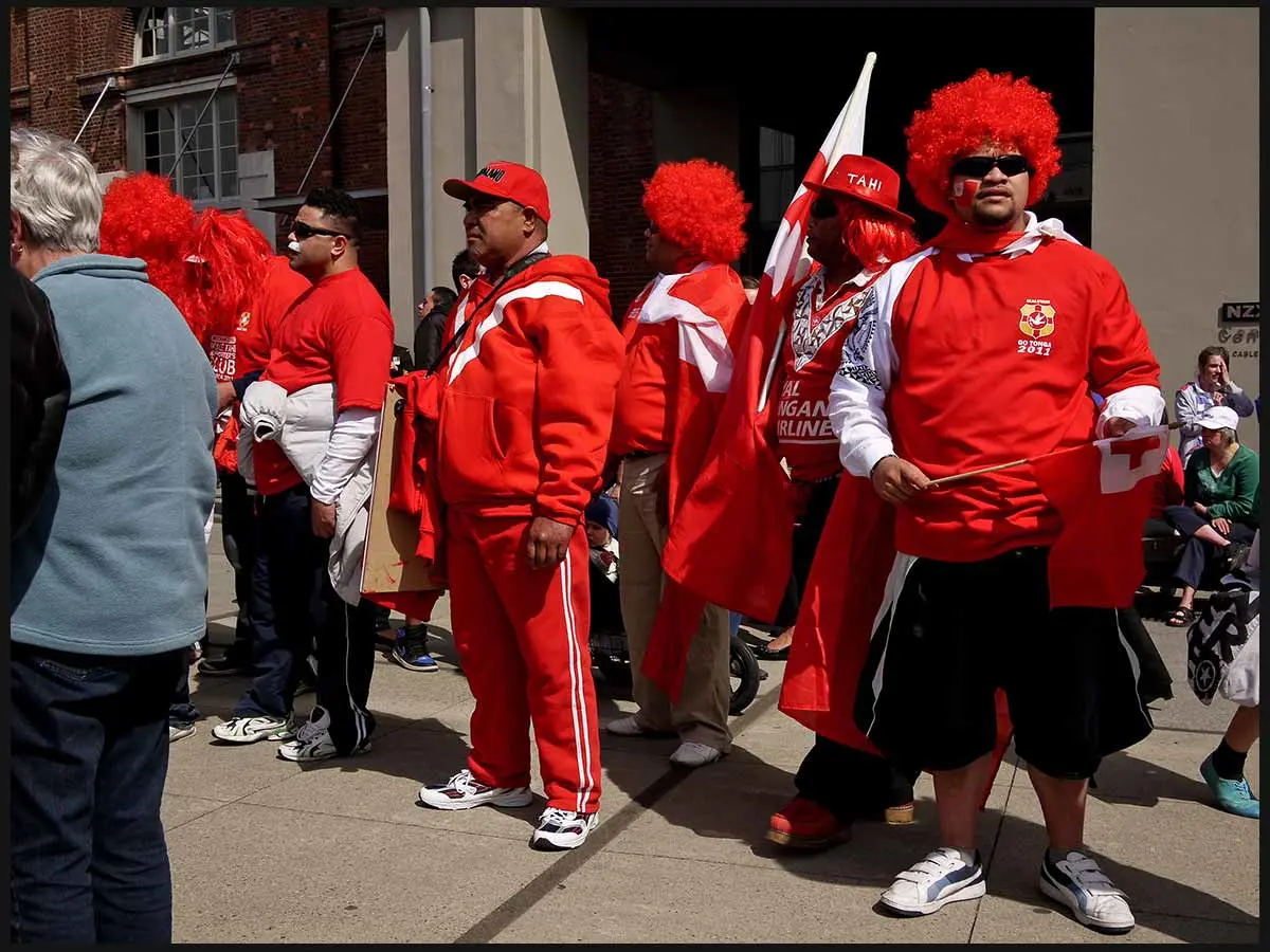 Rugby fans dressed in red wigs and hats to support the Tongan rugby team. Some are holding Tongan flags, and one fan has the flag painted on their face.