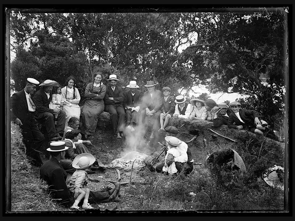 Black and white photo of a large group of adults and children sitting on the grass around a hāngī. They are wearing European-style clothes and hats.