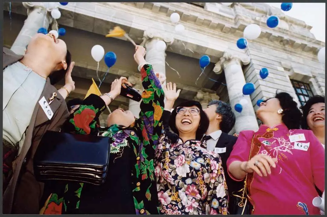 People celebrate on the steps of Parliament holding their hands in the air as blue and white balloons float skyward.