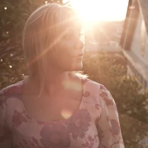 A woman stands in a yard with a house in the background and with the sun setting behind her and causing glare on the camera lens.