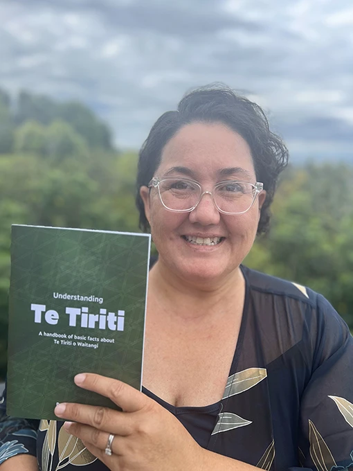 A smiling woman holding a book with a green cover and the text 'Understanding Te Tiriti'.