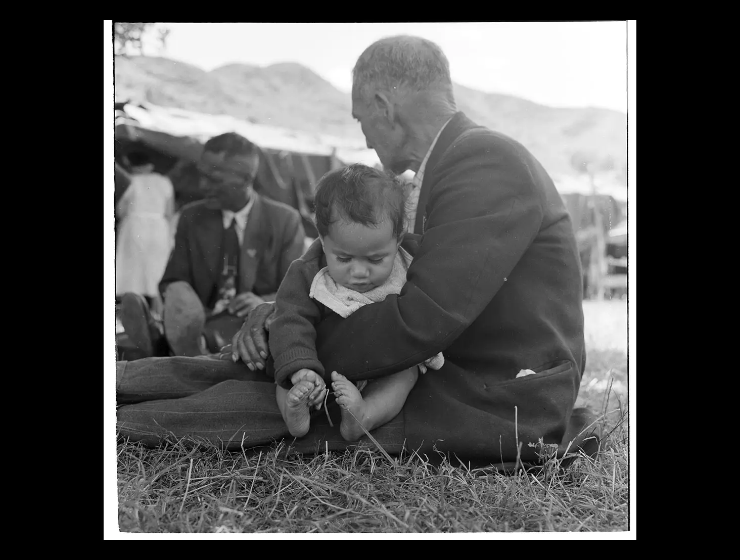 Black and white photo of a koroua (elderly man) sitting on the grass, holding a Māori baby on his lap.