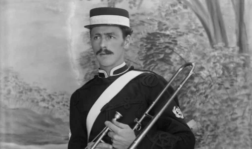 A man in a band uniform and hat holds a trombone with a puzzled look upon his face.