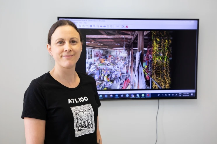 Woman wearing an "ATL100" shirt stands before a large flat screen TV mounted on the wall showing a colour photograph of an interior warehouse/work space.