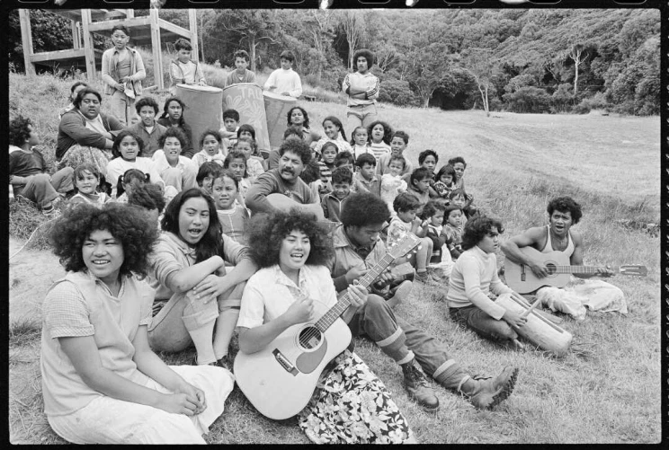 A group of children seated on the ground are singing and two are playing guitar.
