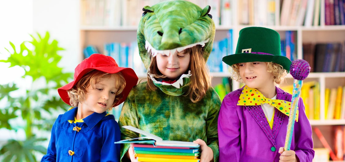 3 children in book character costumes. The middle girl, dressed as an alligator, is holding some books.