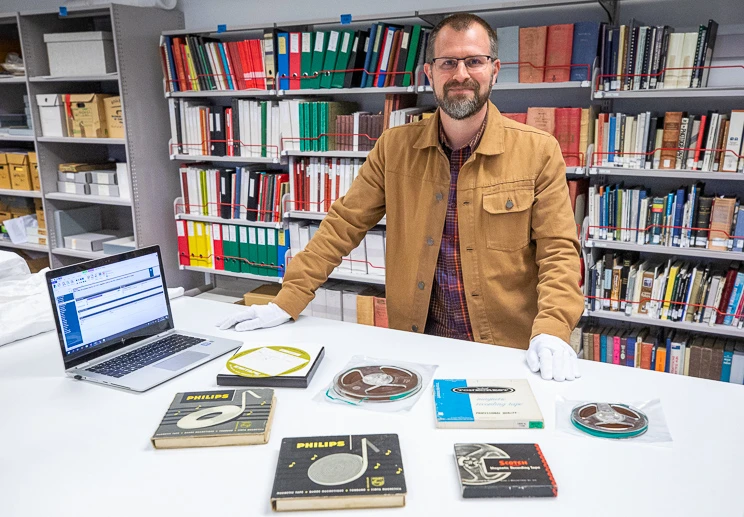 A man standing behind a white table on which sit a variety of collection items including books, open reel tapes and a laptop.