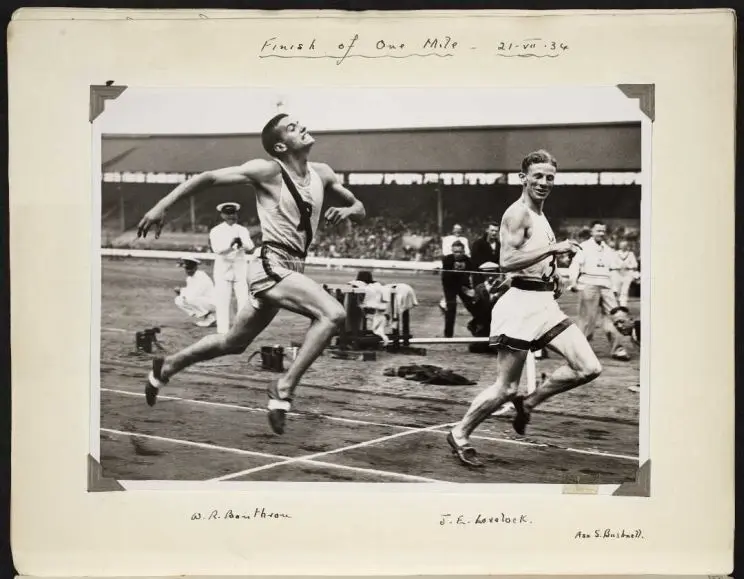 A black and white photo of two men at the finish line with the winner looking relaxed as he crosses and the second place finisher over-exerting himself.