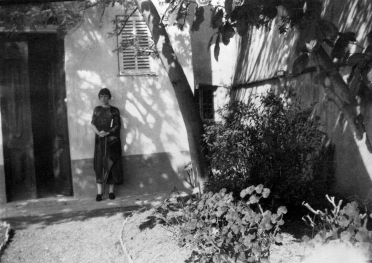 A black and white photo of a woman standing beside the open door to a dwelling with a dirt path and trees and bushes out front.