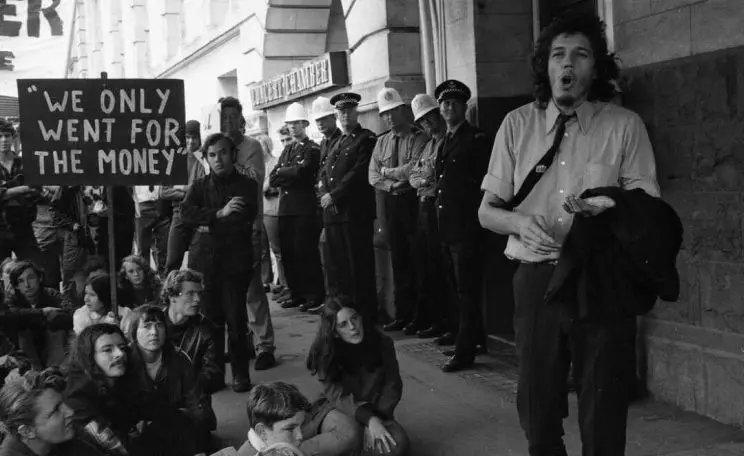 A black and white photo showing protesters seated on the ground with police in the background, and a man standing with his mouth slightly open as if shouting. 