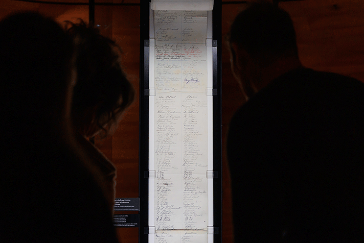 Two silhouetted figures view a framed historical document covered in handwritten signatures, displayed in a dimly lit museum.