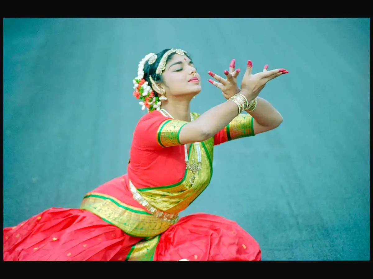 Narmadai Chinniah performing an Indian classical dance, wearing a red, gold and green sari and jewellery.