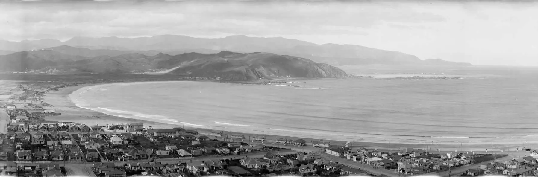 A black and white photo from a ridgetop overlooking the beachside community of Llyal Bay with the large sweep of coast looking out to the headlands and beyond. 