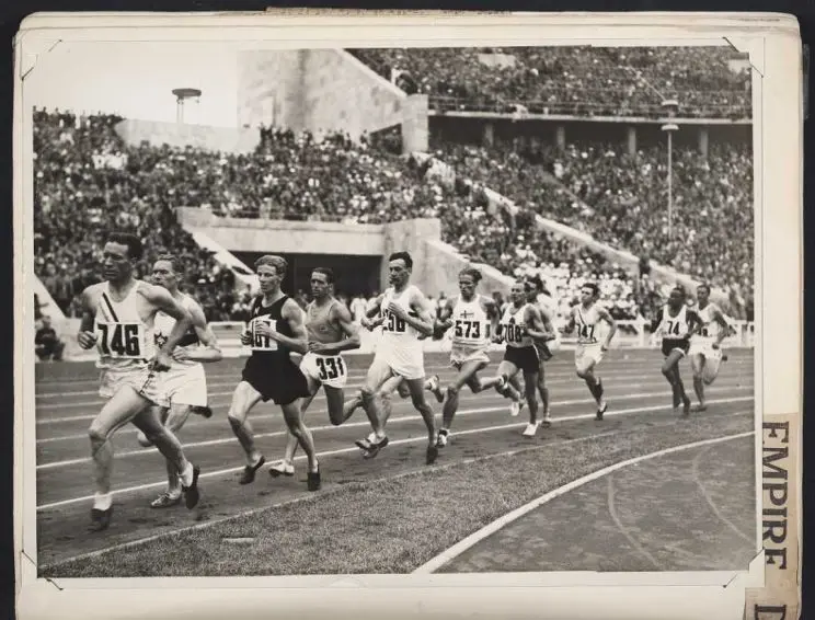 Shows men racing in a 1500 meter with one behind the other as they go around the track, the stands are full in the background.