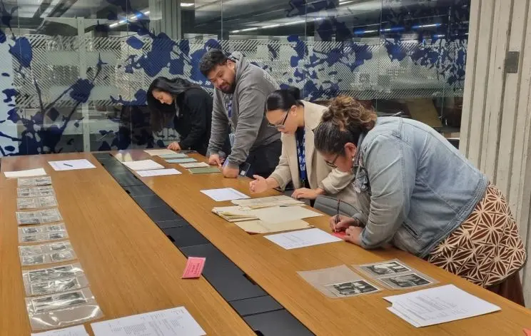 Shows a group of people leaning over a table on which paper documents and black and white photos are displayed. 