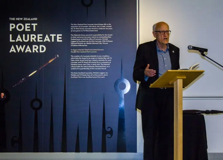 A man stands behind a lectern speaking into a microphone with a banner behind him with the words, in large, white letters: Poet Laureate Award.  