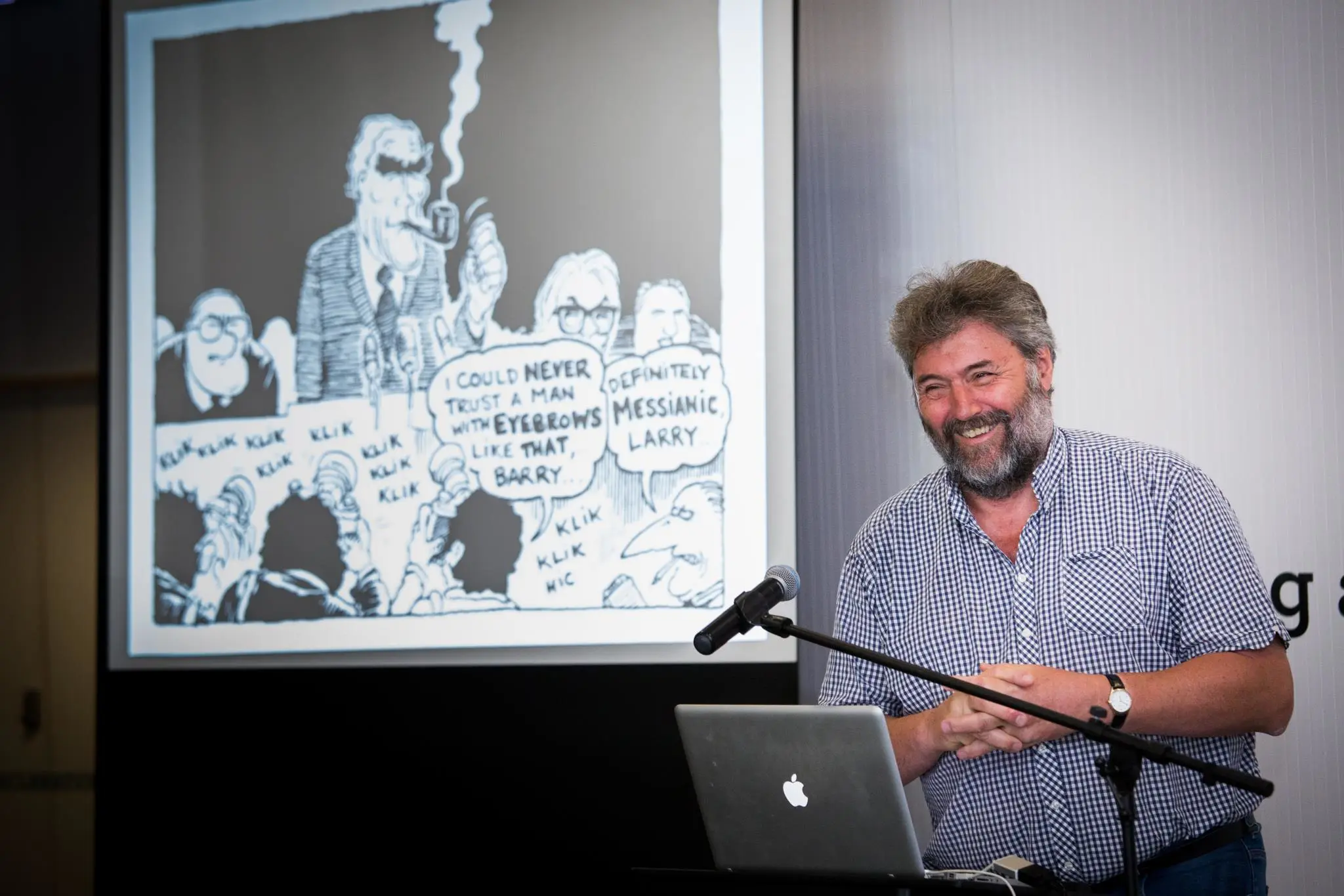 Guardian cartoonist Steve Bell standing behind a lecturn and in front of a screen with a large black and white cartoon on it.