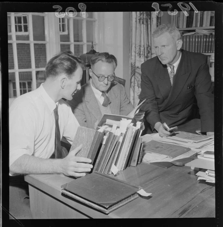 A black and white photo of three men at a desk on which a number of bound volumes are placed and they appear to be all reading something.