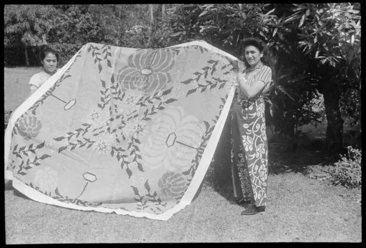 A black and white photo shows two women holding a large tīvaevae between themselves. 