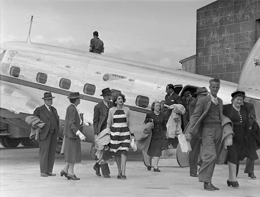 Passengers disembarking onto the tarmac from an airplane with an NAC logo. 