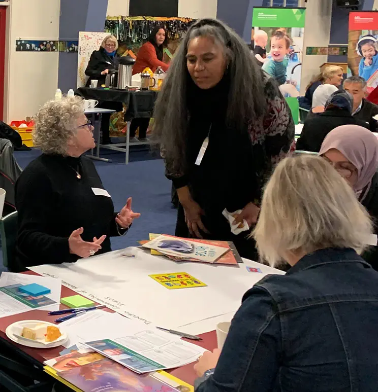 Polynesian woman leaning down and talking to a woman sitting at a low table covered with large and small pieces of paper. 