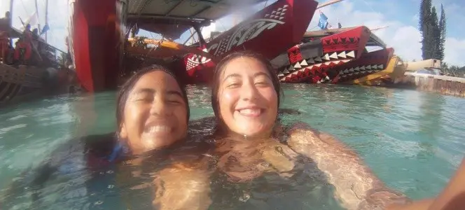 Two girls swimming with painted waka in the background.