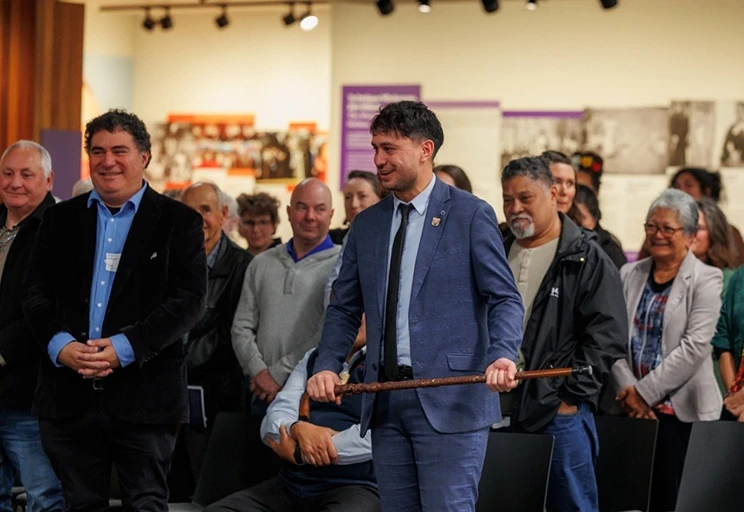 A man in a blue suit holds a carved wooden staff, surrounded by smiling guests in a gallery setting.