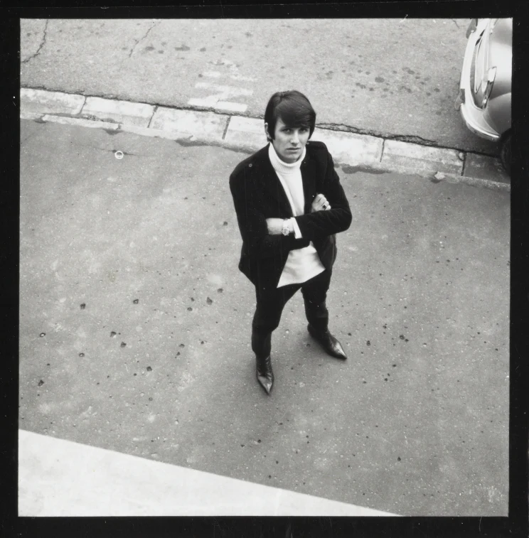A man, viewed from above, is dressed in pointy shoes and a turtleneck stands on the footpath beside a parked car.