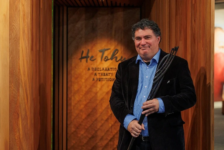 A smiling man in a dark blazer holds a tokotoko outside the He Tohu exhibition, featuring warm wooden panelling.