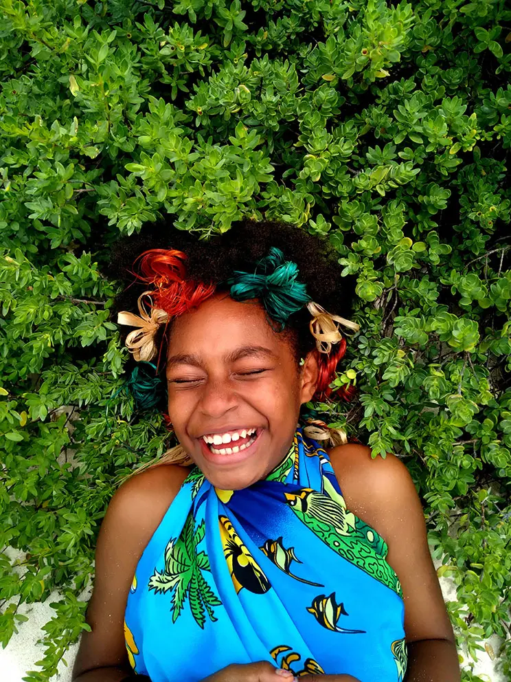 A laughing Pacific woman lying on the ground with green plants all around her. She has colourful ribbons in her hair and a colourful top on. 