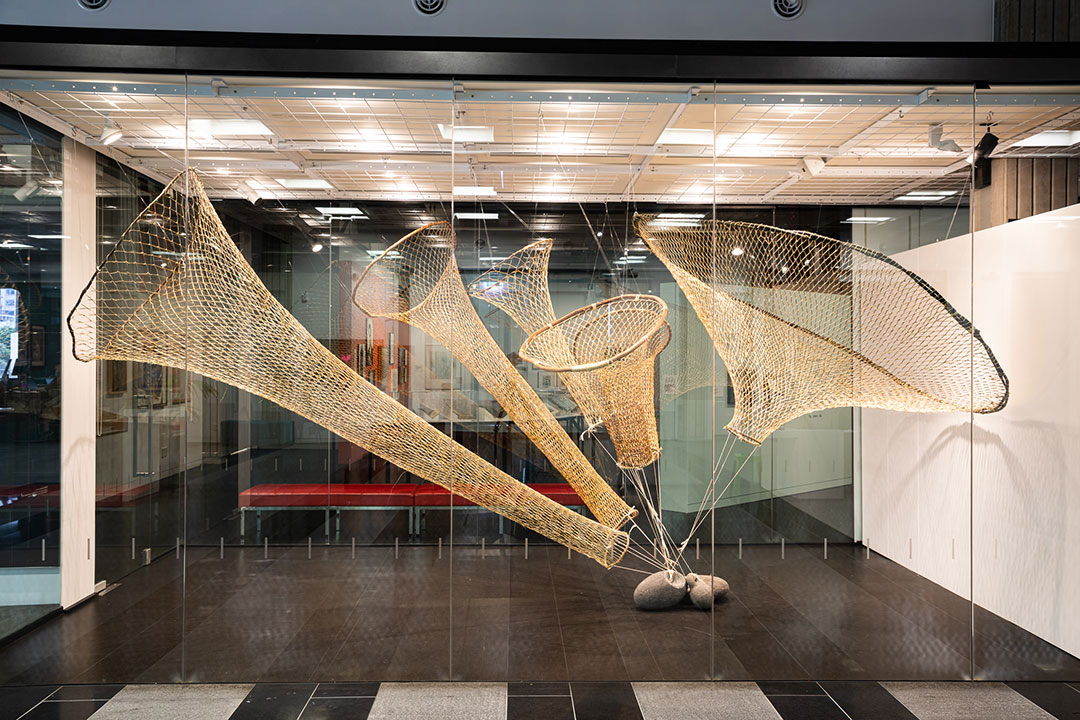5 large eel nets hanging in a large glass box all connected to a stone anchor.