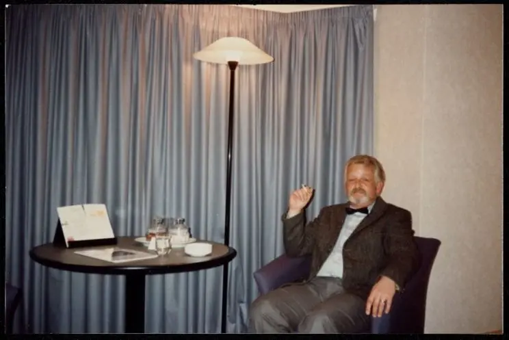 A portrait of a man, seated in an armchair beside a small table and lamp and smoking a cigarette in what appears to be a hotel room. 