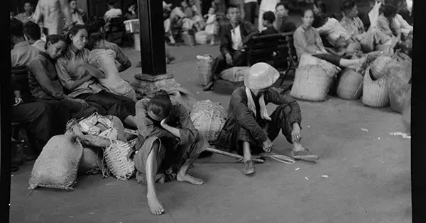 Black and white photo of tired and emaciated-looking Chinese refugees sitting with their sacks of belongings.