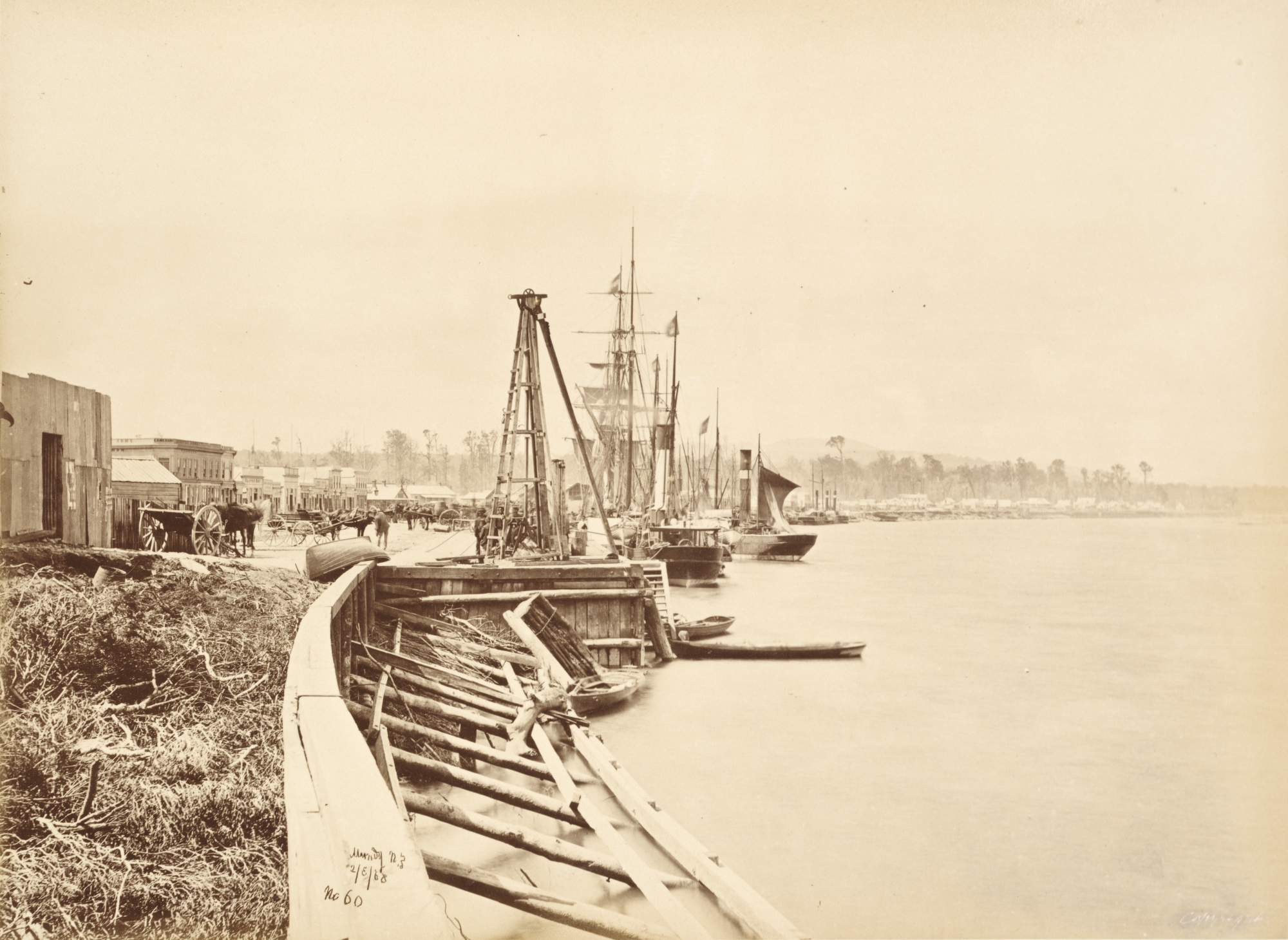 A busy wharf with ships and rowboats lined up and a bustling town at the water's edge.