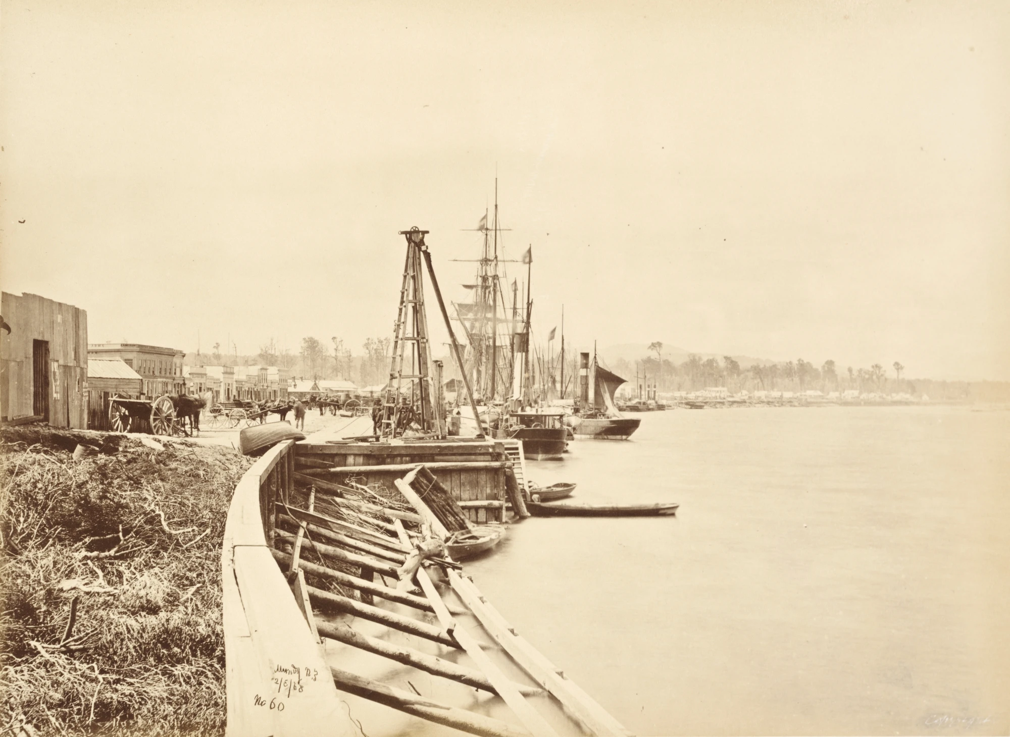 A busy wharf with ships and rowboats lined up and a bustling town at the water's edge. 