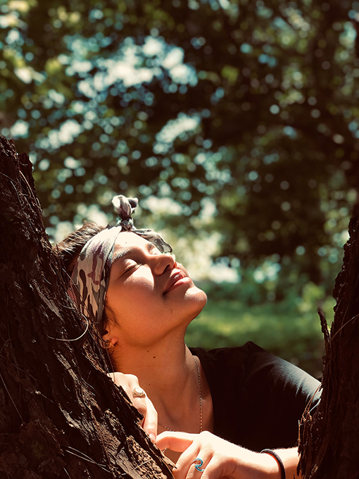 A Polynesian woman standing by a tree with their eyes closed and putting their face towards the sun. 