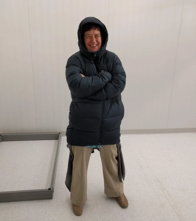 A woman stands smiling in a sterile white room that appears to be very cold as she is wearing a heavy winter parka.