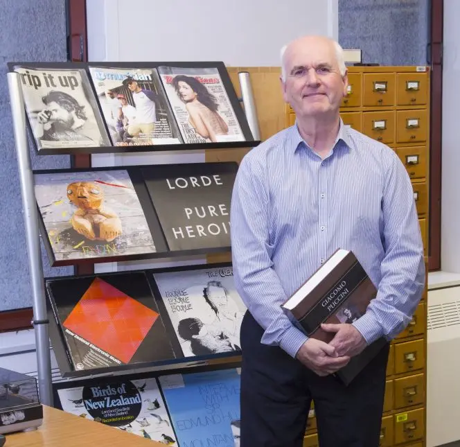 Roger Flury with a copy of his discography on Puccini, in front of several music publications.