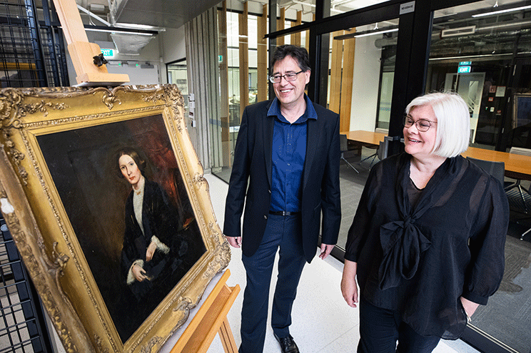 A smiling man and woman looking at a painting of a contemplative woman. The painting is in a heavy gilt frame. 