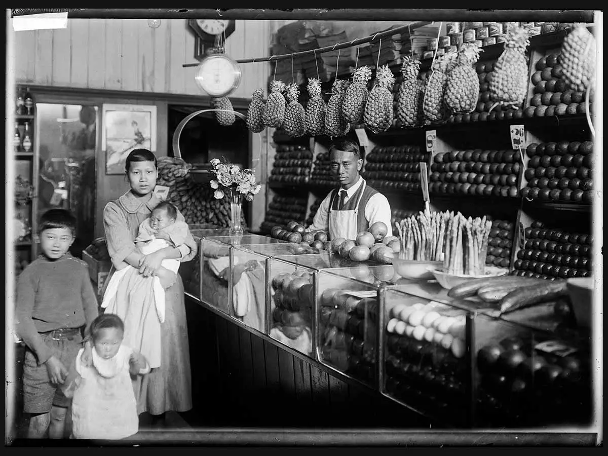 Inside a fruit shop. Shopkeeper, Wong Gar Sui, stands behind a counter with asparagus, apples and cucumbers on top. Above their head hang a row of pineapples. Wong Gar Sui’s wife, Mew Yuen, stands with their 3 children.