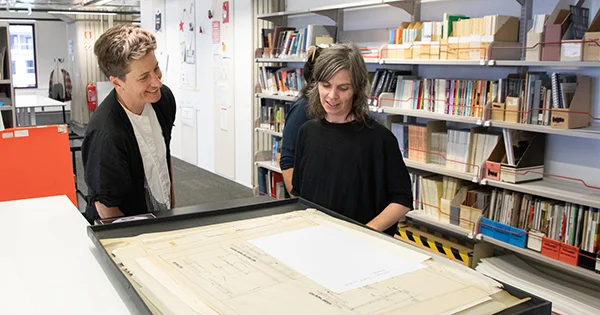 Two people in a room with large bookshelves standing at a work table looking at some papers.