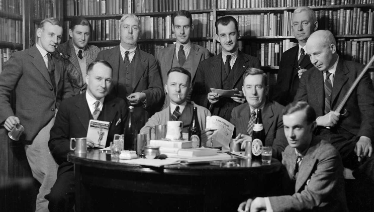 Black and white group photograph of writers arranged around a table with bookshelves in the background. The table has on it tankards, a beer bottle, books and papers.