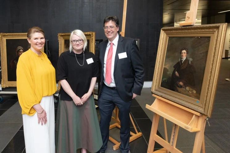 Three smiling colleagues pose for a portrait together while a framed oil painting is to the right of the group.