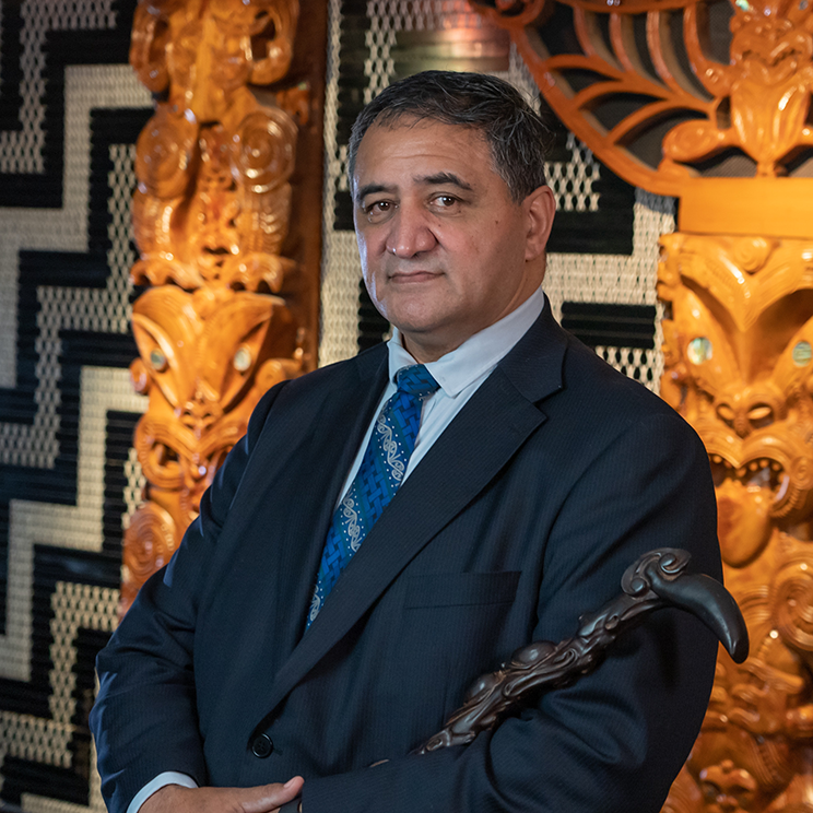 A photo of a man wearing a suit and tie and holding a tokotoko. He stands in front of carved poupou and tukutuku panels.