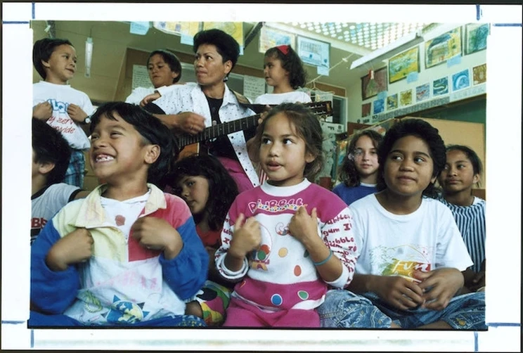 A colour photograph of a classroom setting showing a teacher with guitar surrounded by young children all singing a song together.