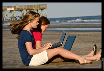 Two girls using laptops on a beach.