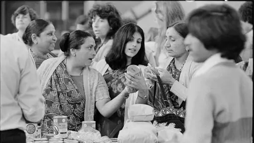 A black and white photo of three women in a market assessing a canned item. 