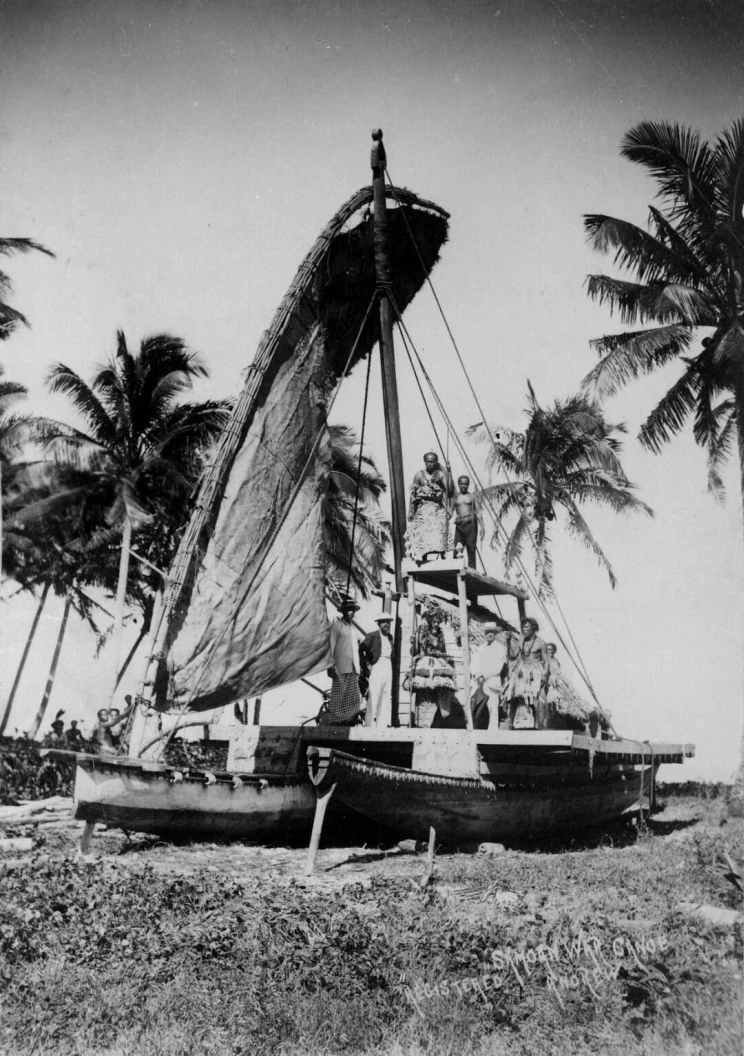 A double-hulled canoe with sail on land is photographed with people standing on it.