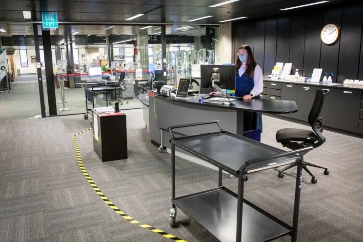 A different view of the woman wearing a mask and standing behind the checkout desk, side angle shows a yellow and black stripped tape on the ground indicating where to stand to ensure proper distancing between staff and client.