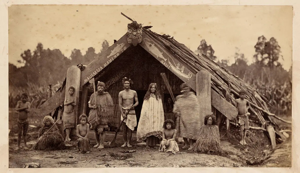 Black and white photograph of a Māori group in front of a wharepuni. 'Maui Mua Maui Roto' is written on the maihi.